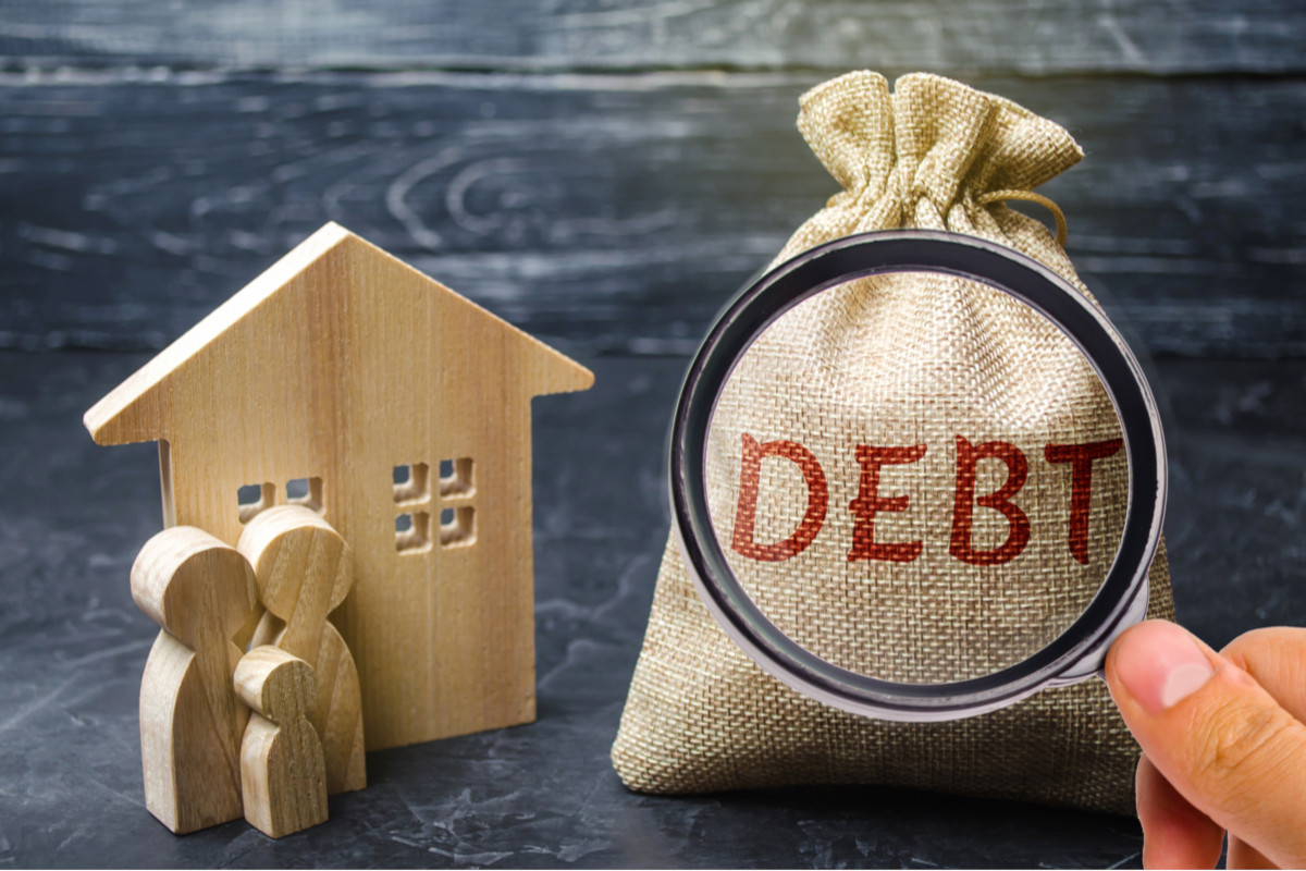 A wooden block house with a bag of money with the word "debt" on it. 