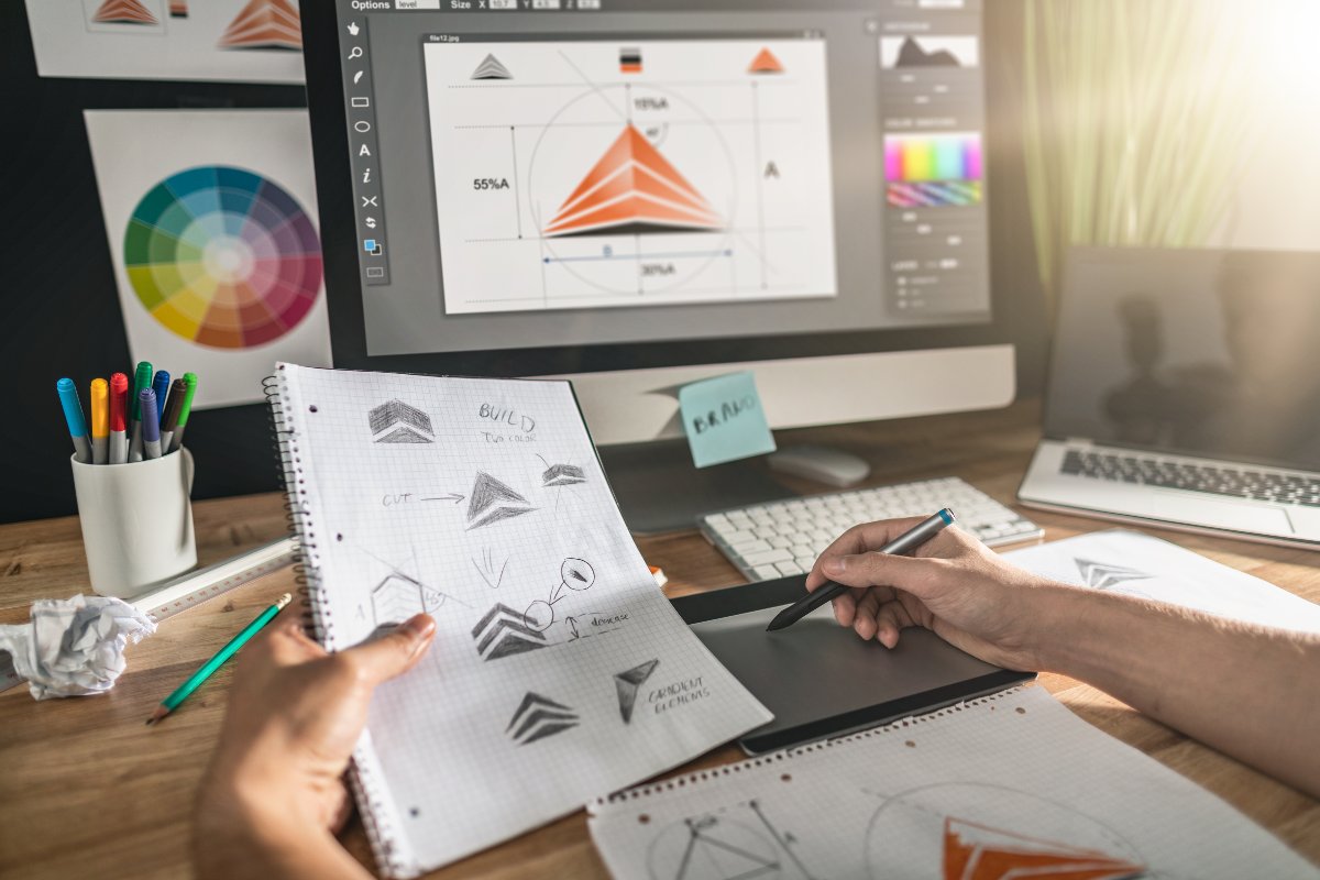 Graphic designer holding notebook in one hand and drawing logo on computer with the other hand.