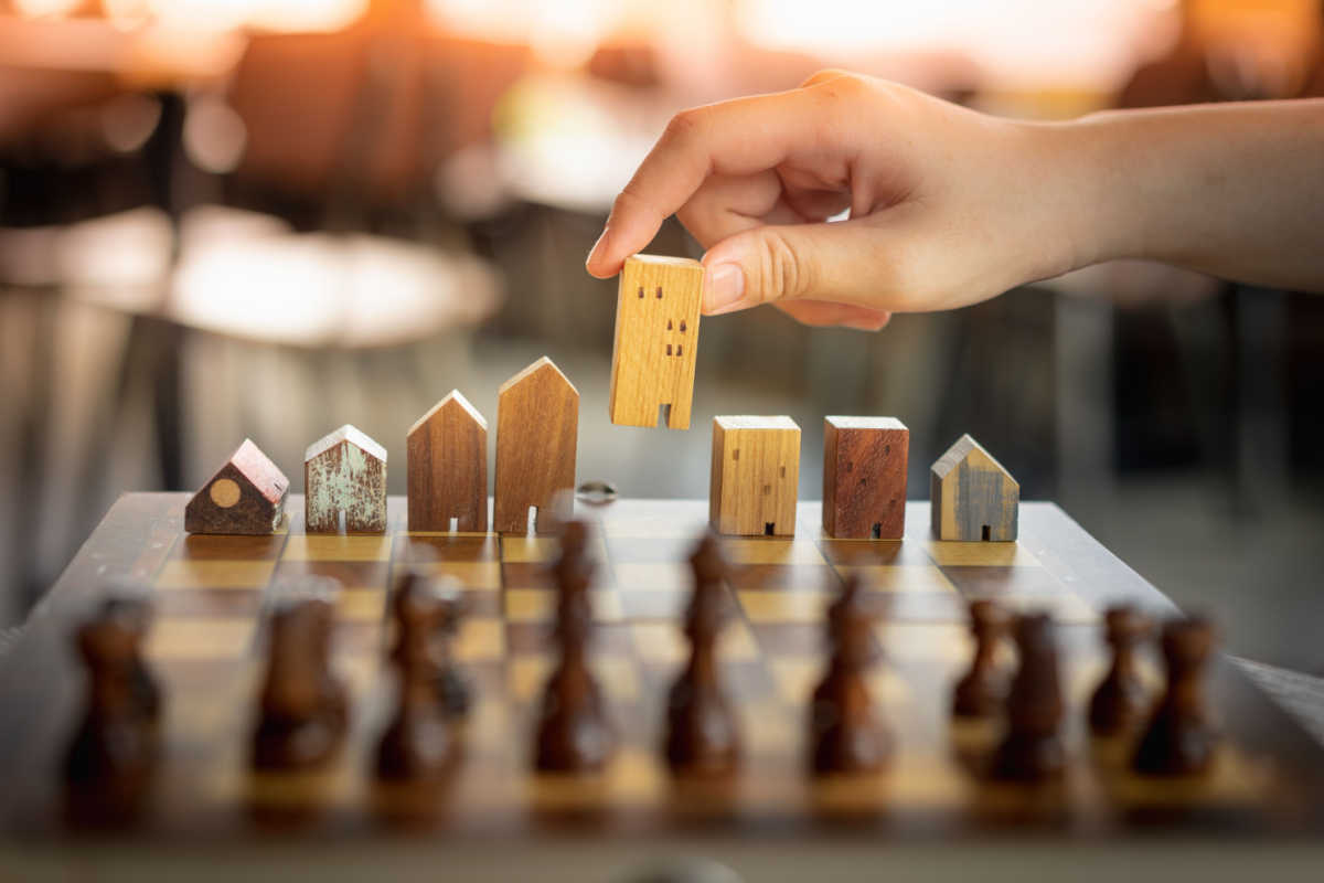 A set of wooden house and building figurines on top of a chess table. A hand is picking up a building figurine.
