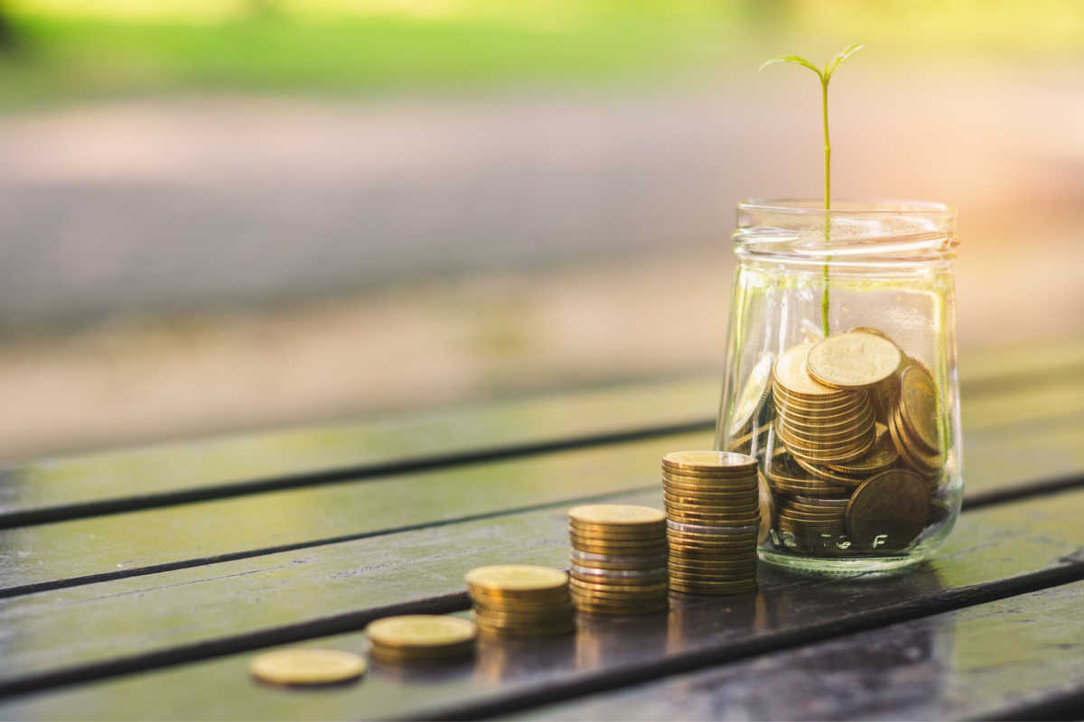 golden coins stack on wood table and coin in glass jar green leaves growth on with blur nature